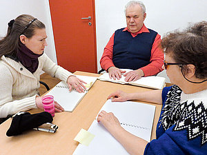 Kursleiter und zwei Schülerinnen sitzen an einem Tisch und betasten Seiten in Blindenschriftbüchern