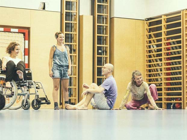Mehrere Menschen, eine davon im Rollstuhl, sitzen oder stehen lächelnd in einer Turnhalle.