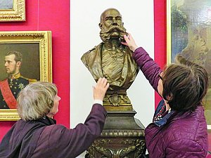 Zwei ältere Frauen von hinten ertasten eine goldene Büste eines Kaisers