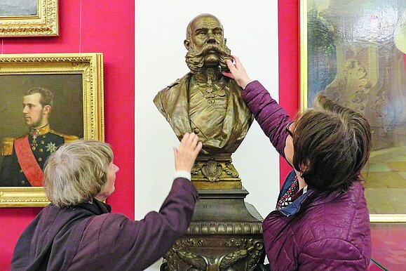 Zwei ältere Frauen von hinten ertasten eine goldene Büste eines Kaisers