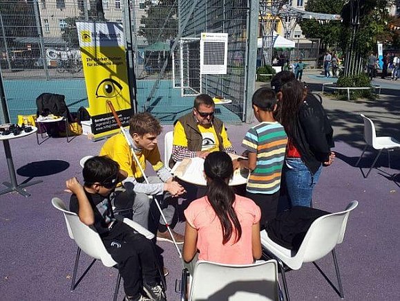 An einem kleinen runden Tisch sitzen zwei sehbeeinträchtigte Männer und vier Kinder, die ihnen zuhören. 