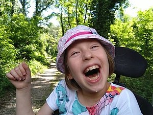 Die mehrfach behinderte Anna sitzt in ihrem Rollstuhl im Grünen und freut sich mit einem breiten, ansteckendem Lächeln