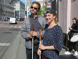 Eine Frau und ein Mann stehen mit je einem Langstock in der Hand bei einer Straßenbahnstation. 