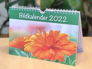 Ein Stehkalender mit Blumenmotiv und Aufschrift "Bildkalender 2022" auf der Vorderseite steht auf einem Tisch.