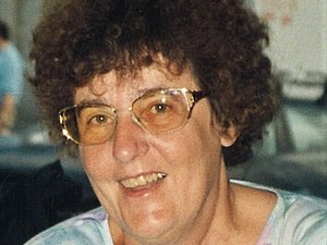 Portrait einer Dame mittleren Alters mit braunen Locken und Brille, freundlich lachend.