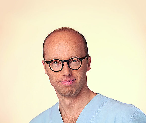 Ein Mann mit Brille und blauem Arztgewand lächelt in die Kamera