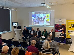 Sechs Personen sitzen bei einer Podiumsdiskussion vor Publikum. Der Mann links spricht in ein Mikrofon. Hinter dem Podium wird eine Liveübertragung der Veranstaltung an die Wand projiziert. Daneben stehen Banner von VIDEBIS und der Hilfsgemeinschaft der Blinden und Sehschwachen