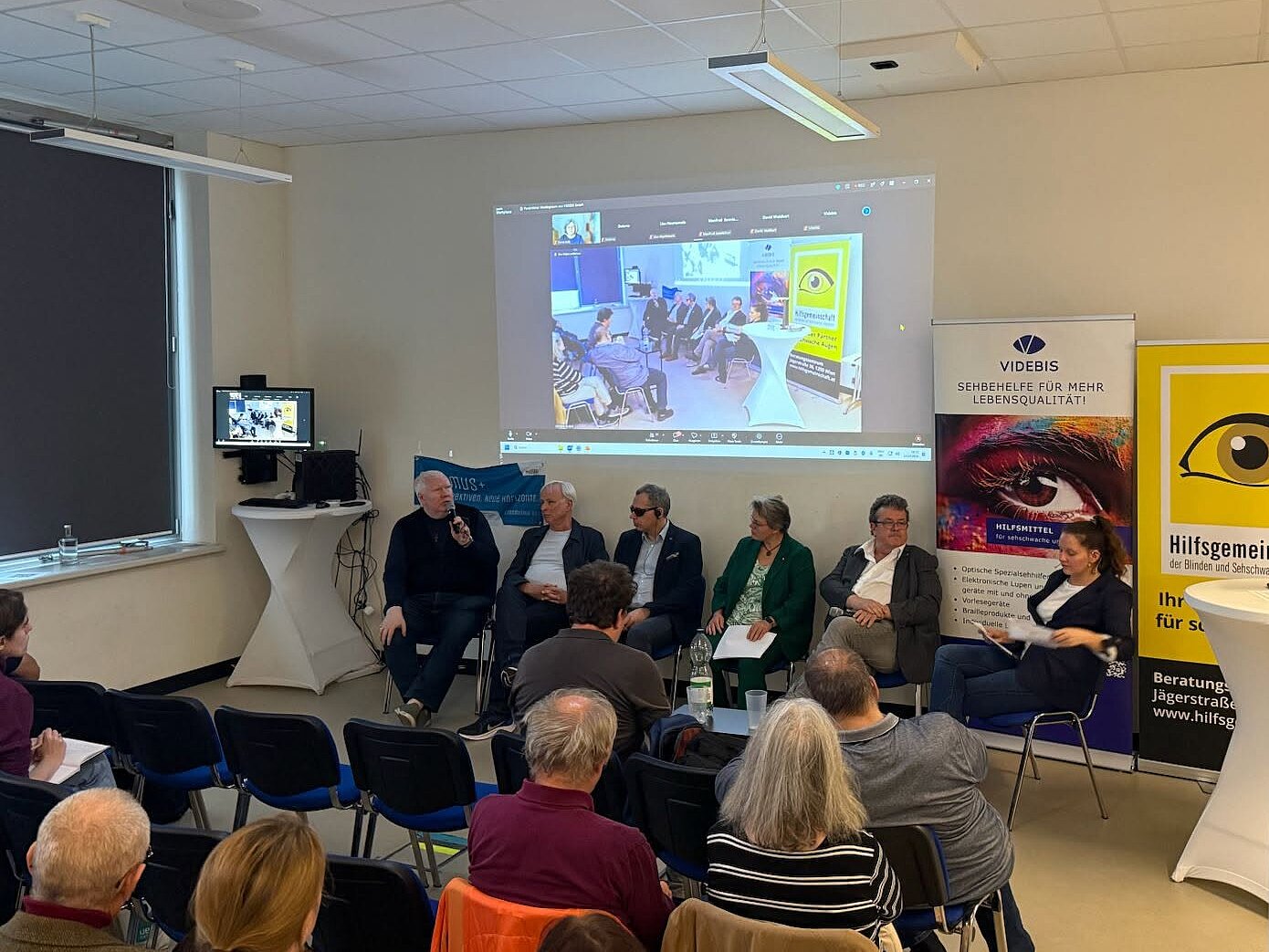 Sechs Personen sitzen bei einer Podiumsdiskussion vor Publikum. Der Mann links spricht in ein Mikrofon. Hinter dem Podium wird eine Liveübertragung der Veranstaltung an die Wand projiziert. Daneben stehen Banner von VIDEBIS und der Hilfsgemeinschaft der Blinden und Sehschwachen