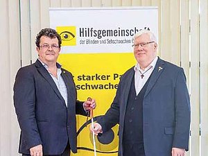 Zwei Männer mit Anzug vor Hilfsgemeinschaft Roll-Up, halten beide weißen Stock