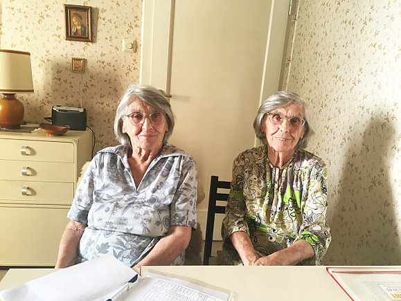 Zwei Zwillinge mit Brille und grauen Haaren sitzen nebeneinander an einem Tisch.