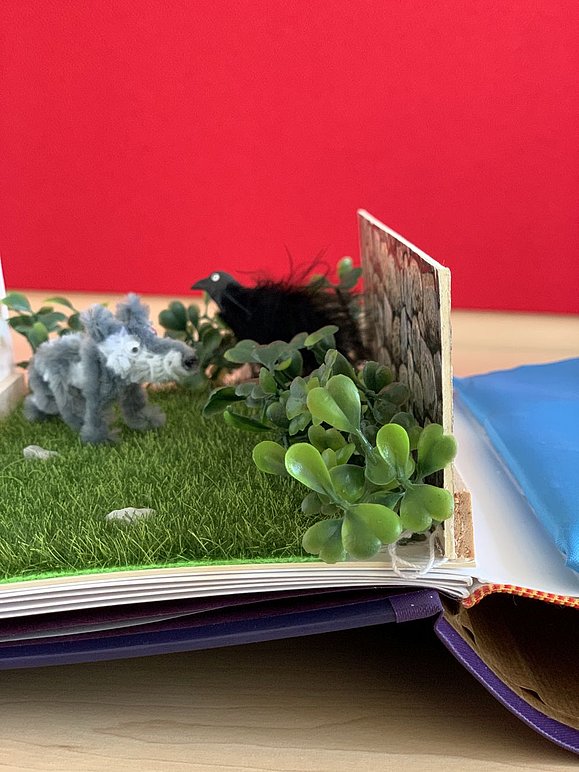 Seite im Buch mit hervorstehendem Gras, flauschiger Wolfsfigur und schwarzem Rabe mit Drahtfüßen