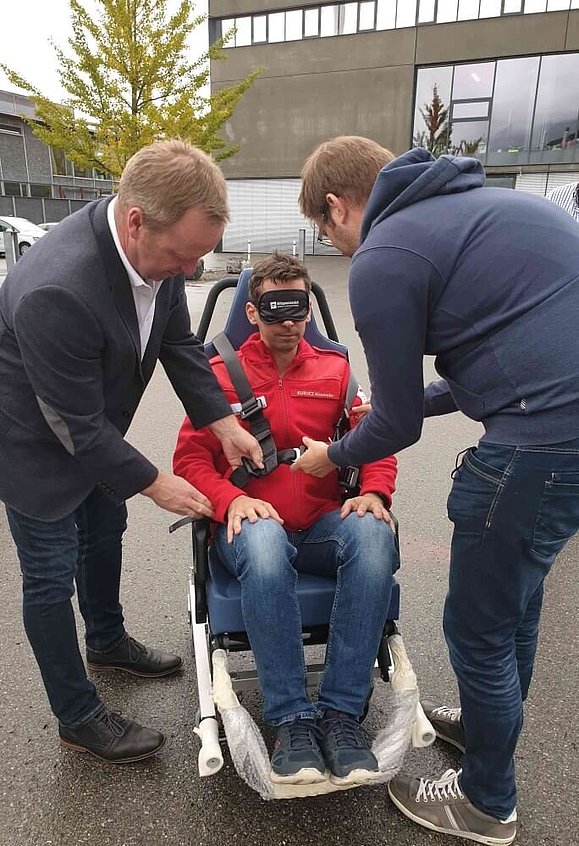 Ein Mann mit Augenbinde sitzt auf einem Rettungsstuhl, zwei Männer stehen links und rechts davon und helfen ihm mit den Gurten