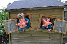 Zwei Kinder spielen mit dem Kuscheltier-Seestern und schauen aus den Fenstern eines Holzhauses