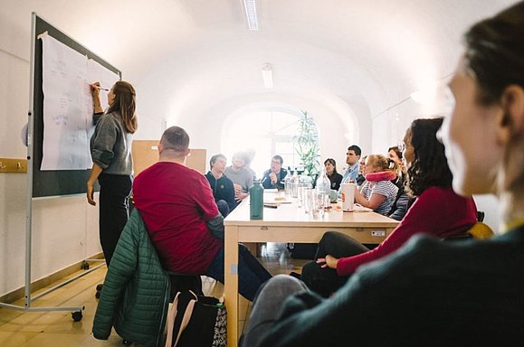 junge Frau schreibt an Tafel, mehrere junge Menschen an großem Tisch sitzend schauen an die Tafel