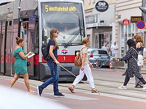 Personen kreuzen an Zebrastreifen die Straße, auch eine junge Frau mit Langstock, sunkler Brille und Rucksack. Dahinter wartet eine Straßenbahn mit der Aufschrift: "5, Westbahnhof"