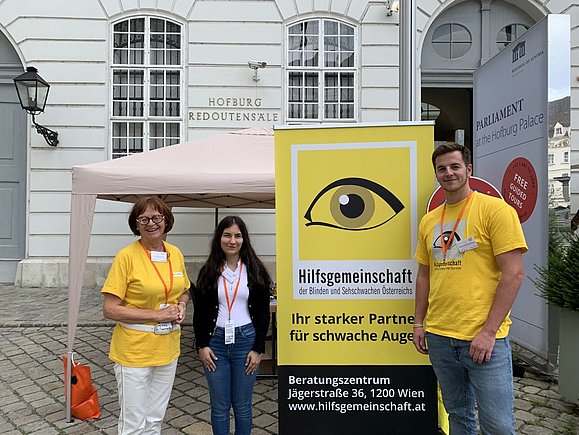 Drei Leute neben gelbem Roll-Up mit Auge und Logo Hilfsgemeinschaft der Blinden und Sehschwachen Österreichs.