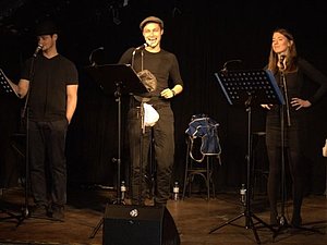 Zwei Männer und eine Frau stehen auf einer Bühne, vor ihnen Notenständer und Mikrofone. 