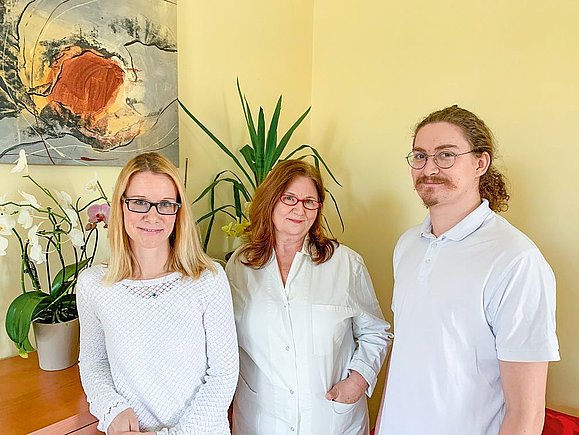 Zwei Frauen und ein Mann in weißer Kleidung stehen nebeneinander in einem hellen Raum.