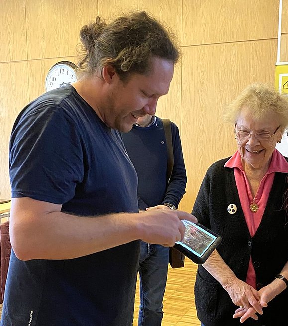 Mann mit Pferdeschwanz zeigt etwas auf dem Tablet, rechts von ihm eine ältere Dame mit Blinden-Anstecker mit begeistertem Gesichtsausdruck