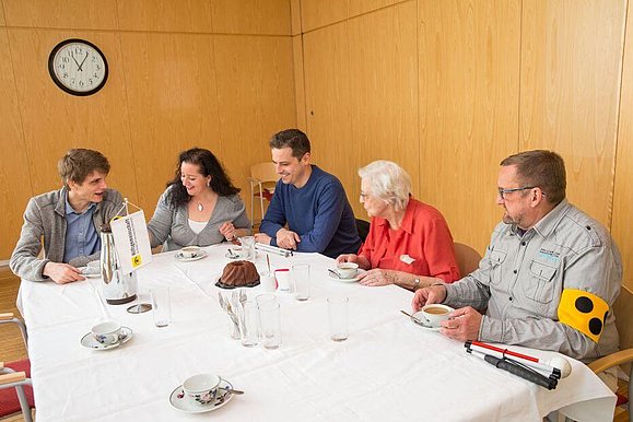 5 Menschen sitzen um einen gedeckten Tisch beisammen und tauschen sich aus.