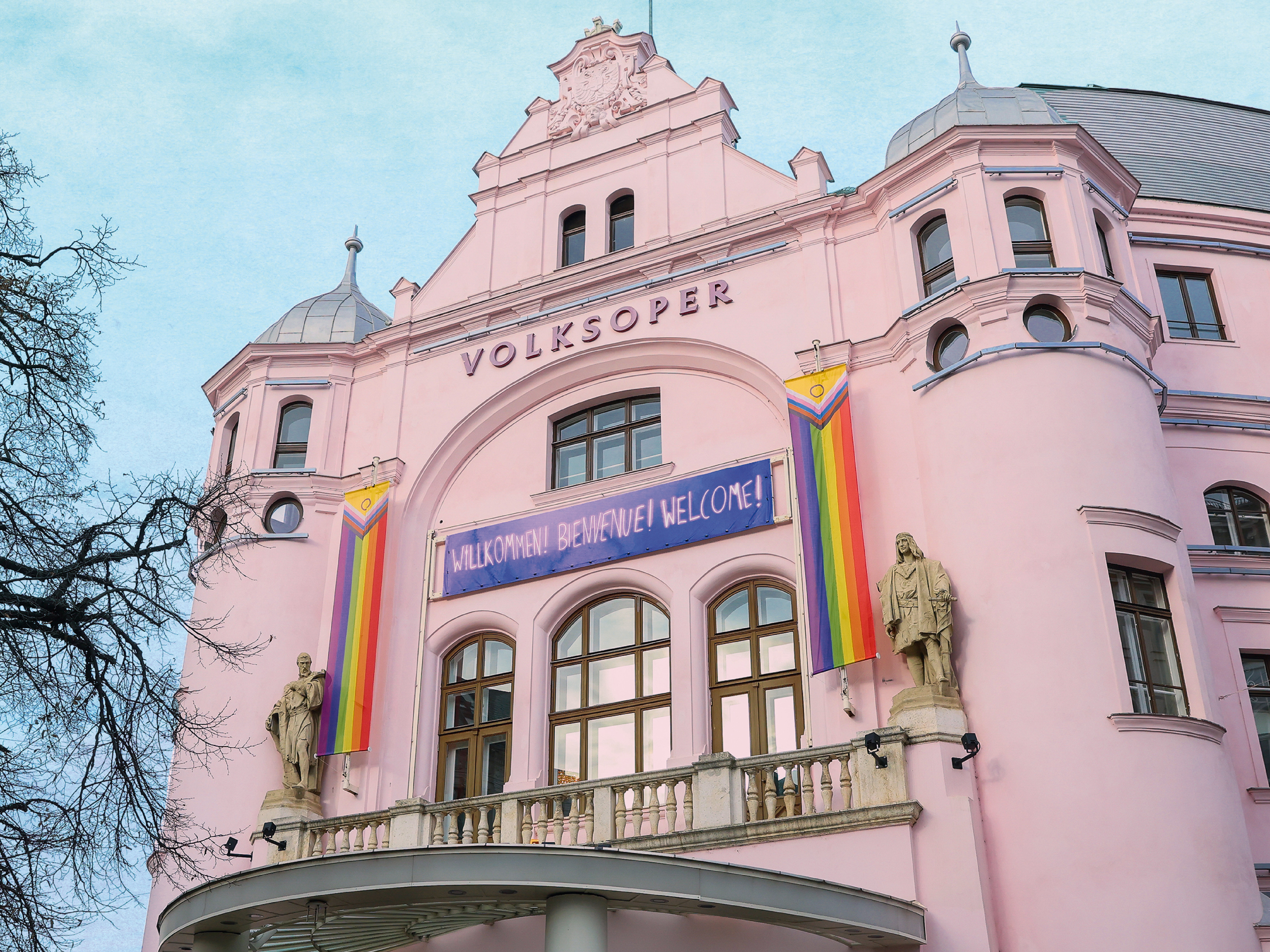 Rosa historisches Gebäude mit der Aufschrift „Volksoper“, geschmückt mit Regenbogenfahnen und einem Banner über dem Eingangsbereich.