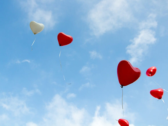 Rote und weiße Herzluftballons fliegen in einem blauen Himmel mit kleinen, weißen Wölkchen.