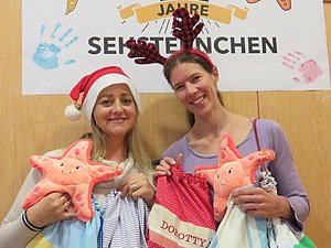 Zwei Mitarbeiterinnen der Hilfsgemeinschaft zeigen Kuschelseesterne und Turnbeutel mit Namen drauf her. Dabei Tragen sie Weihnachtsmützen.