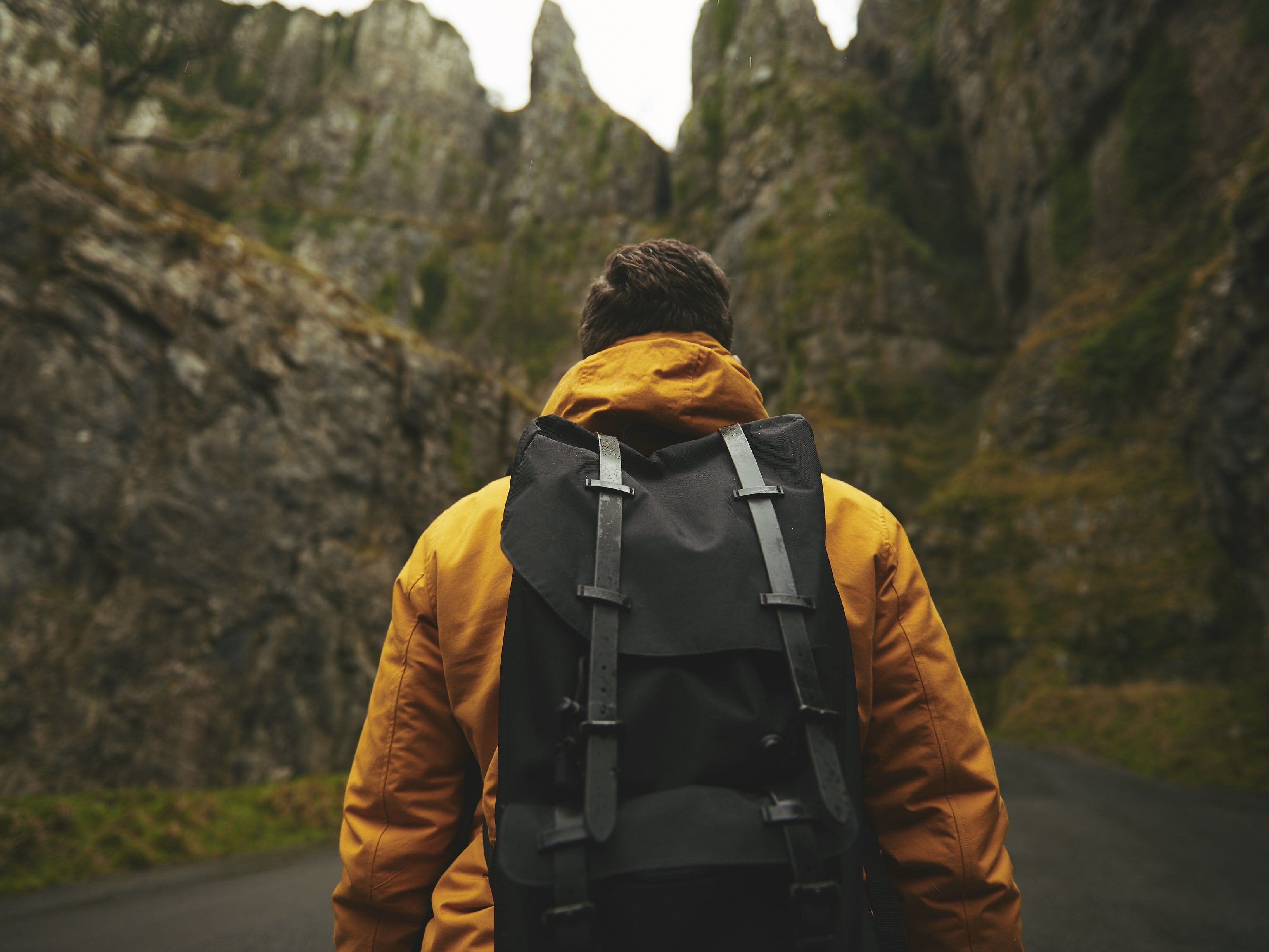 Person mit gelber Outdoorjacke und schwarzem Rucksack steht auf einer Straße zwischen steilen, moosbewachsenen Felswänden.