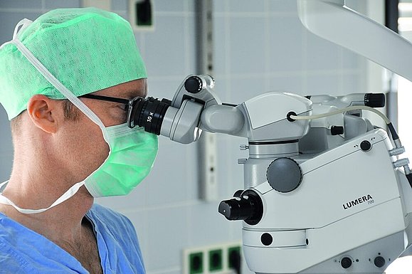 Ein Mann mit OP-Haube und Mundschutz schaut während einer Augen-OP durch ein Augen-OP-Gerät