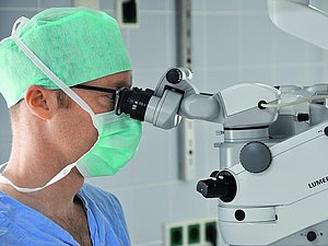 Ein Mann mit OP-Haube und Mundschutz schaut während einer Augen-OP durch ein Augen-OP-Gerät