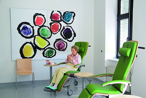 Eine ältere Frau sitzt auf einem grünen Sessel und isst ein Joghurt, neben ihr steht ein weitere Sessel und ein kleiner Tisch, dahinter hängt ein großes Bild mit bunten Kreisen