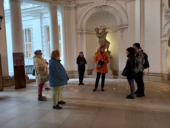 mehrere Menschen teils mit Langstock stehen im Kreis vor Skulptur in Arkadenbogen