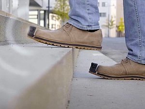 Person mit braunen InnoMake-Schuhen geht Stufen aufwärts. InnoMake-Sensor sendet Wellen aus.