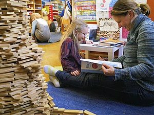 Blick in einen mit vielen bunten Materialien ausgestatteten Fachraum in dem eine Lehrerin und eine Volksschülerin lesend am Boden sitzen