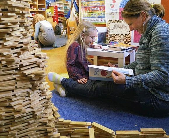 Blick in einen mit vielen bunten Materialien ausgestatteten Fachraum in dem eine Lehrerin und eine Volksschülerin lesend am Boden sitzen