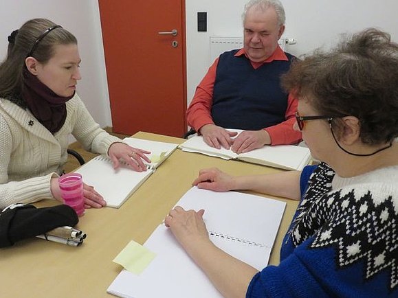 An einem Tisch sitzen drei blinde Menschen vor ihren Braille-Büchern.