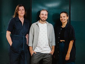 drei Personen vor grüner Wand, junge Frau mit schwarzem Jumpsuit und langen braunen Haaren, junger blonder Mann mit Bart und Hemd mit weißem Shirt, junge Frau mit zusammengebundenen Haaren und schwarzem Hemd und jeans lächeln