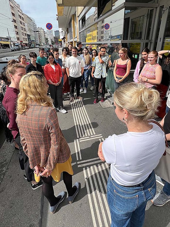 Eine Gruppe junger Menschen steht auf einem Gehweg in der Stadt vor einem Gebäude und hört zwei Frauen zu, die offenbar etwas erklären. Auf dem Boden verlaufen taktile Leitlinien für sehbehinderte Menschen. Im Hintergrund sind Geschäfte, Autos und ein Straßenzug zu sehen.