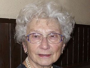 Eine ältere Dame mit Dauerwelle, großer Brille und mehrerer Ketten um den Hals blickt leicht lächelnd in die Kamera