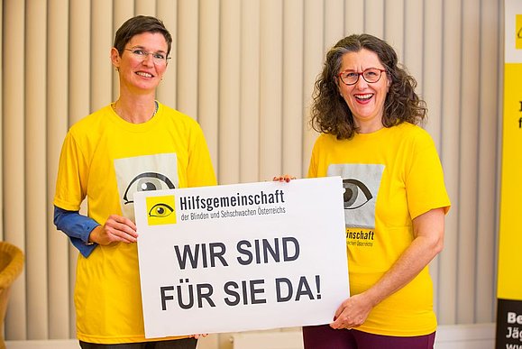 Zwei Frauen mit gelbem T-Shirt halten Schild mit der Aufschrift: Wir sind für Sie da!