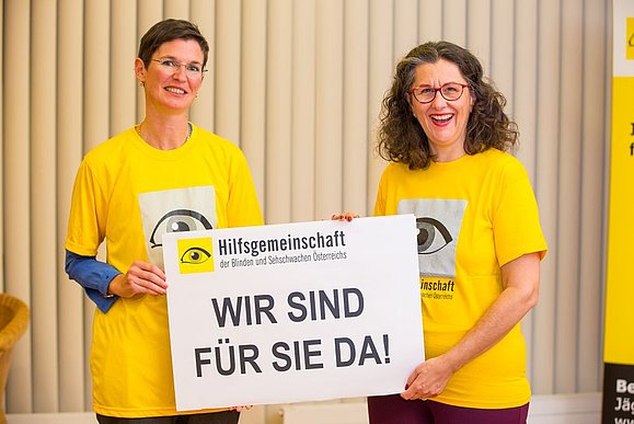 Unsere Mitarbeiterinnen der Testamentsabteilung informieren rund um das Thema Testamente und Vorsorge. Zwei Frauen mit gelbem T-Shirt halten Schild mit der Aufschrift: Wir sind für Sie da!