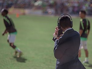 Mann mit Anzug von hinten steht am Rande von Fußballfeld und hält Mikrofon und Kopfhörer