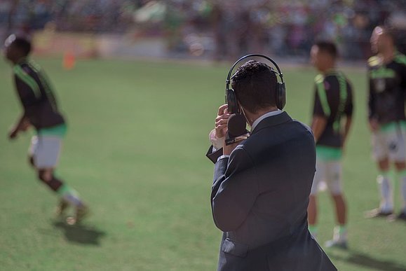 Mann mit Anzug von hinten steht am Rande von Fußballfeld und hält Mikrofon und Kopfhörer