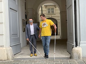 Mann mit gelbem Shirt führt man mit Augenbinde und weißem Stock