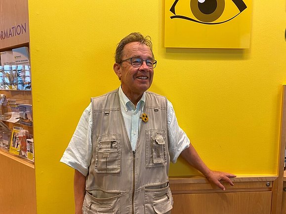 Ein älterer Herr mit Anstecknadel und Brille steht fröhlich lächelnd vor dem Logo der Hilfsgemeinschaft. 