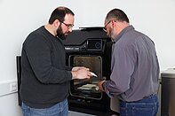 Incubation_Center_3D_Drucker__c_Schedl_Hilfsgemeinschaft_A5.jpg Zwei Männer begutachten einen Gegenstand aus dem 3D Drucker, der hinter ihnen steht