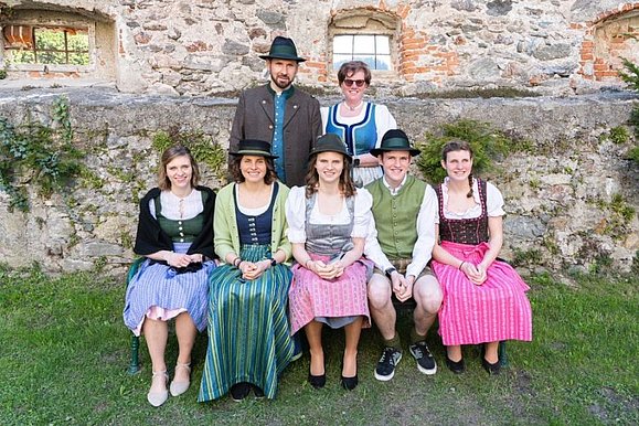 Gruppenfoto mit 2 Reihen, alle in Tracht: hinten steht Mann und Frau, vorne sitzen 5 junge Menschen. im Hintergrund Steinmauern