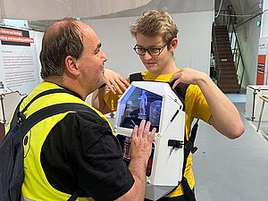 Junger Mann hat einen Hartschalenrucksack mit Sichtfenster vor den Bauch geschnallt. Mann mit Sehbehinderung tastet ab.