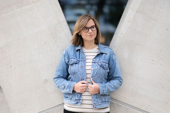 Junge Frau mit schulterlangen dunkelblonden Haaren und Brille steht vor Betonwand und hält ihre Hände an Jeansjacke fest