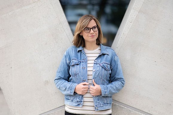 Junge Frau mit schulterlangen dunkelblonden Haaren und Brille steht vor Betonwand und hält ihre Hände an Jeansjacke fest